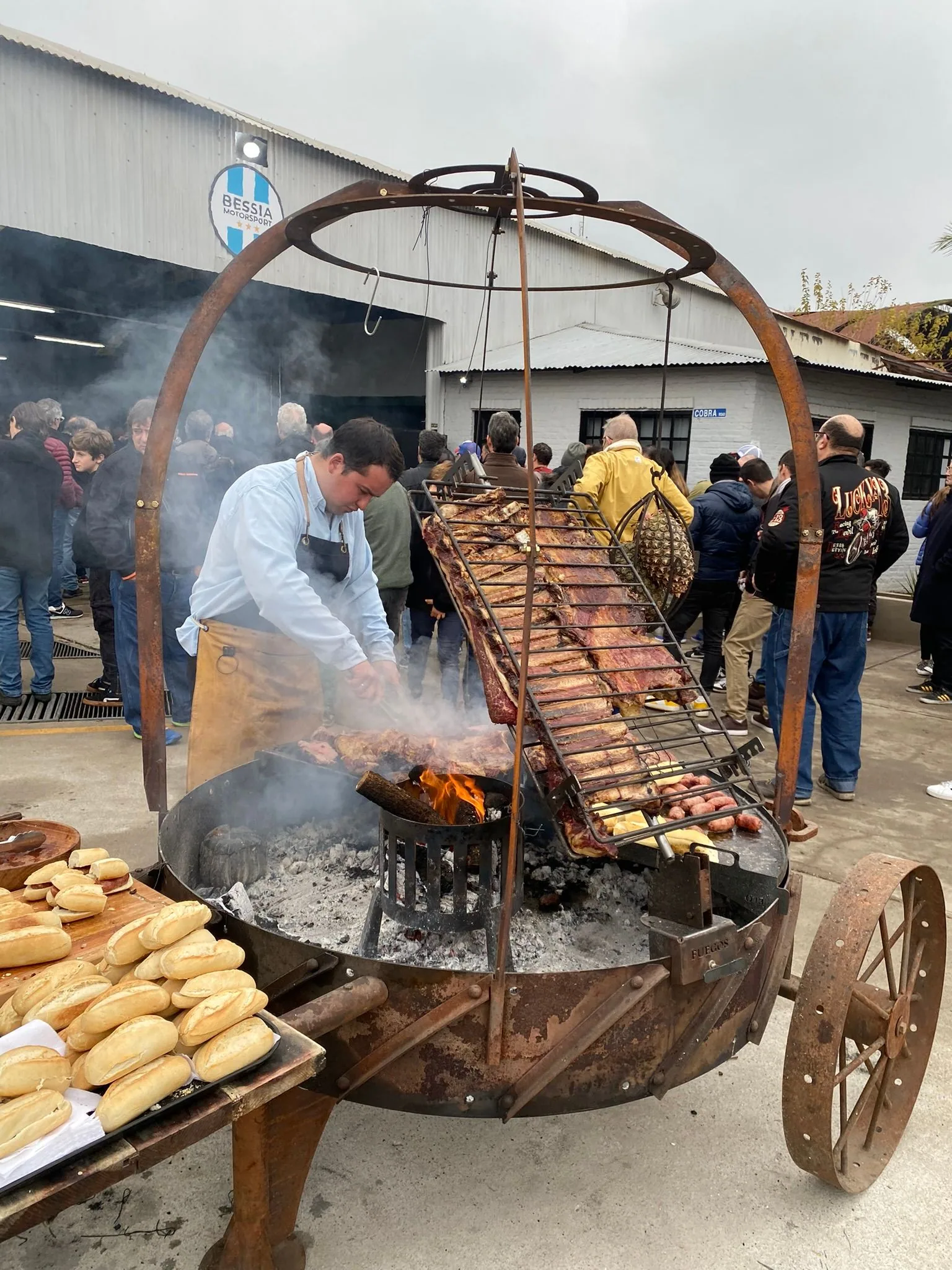 A chef working the round grill loaded with meat and corn, guests behind